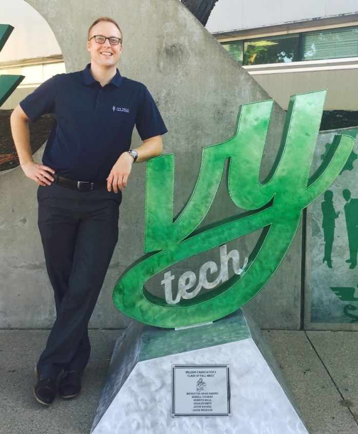 Andrew Bourne, Assistant Director of Early College Programming & Coordinator of the Ivy Career Advancement Program with “I in Ivy” sculpture near Student Life Center entrance. Sculpture was made by the fall welding fabrication II class, 2015.